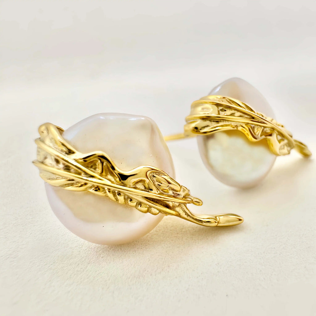 A pair of exquisite earrings featuring large baroque pearls, each embraced by a detailed gold setting with a dynamic, flowing design that evokes a sense of movement. The golden elements wrap around the pearls, highlighting their natural beauty and irregular shapes, set against a light background that enhances the jewelry's luxurious feel.