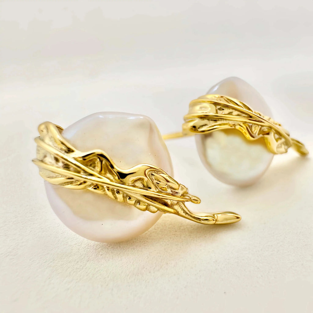 A pair of exquisite earrings featuring large baroque pearls, each embraced by a detailed gold setting with a dynamic, flowing design that evokes a sense of movement. The golden elements wrap around the pearls, highlighting their natural beauty and irregular shapes, set against a light background that enhances the jewelry's luxurious feel.