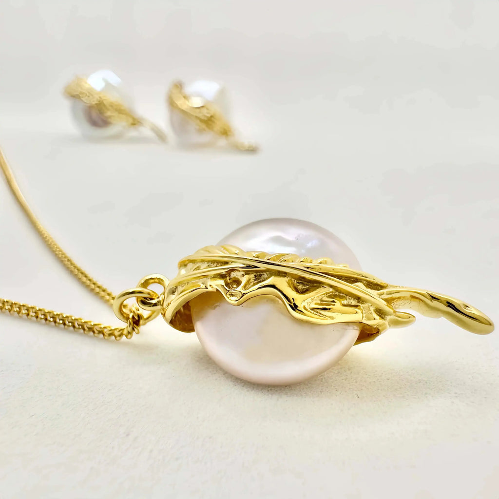 A striking pendant featuring a large, baroque pearl elegantly encased in a flowing, gold metal design reminiscent of feathers or foliage. This artistic wrap is connected to a fine gold chain, creating a luxurious and eye-catching piece of jewelry against a white background.