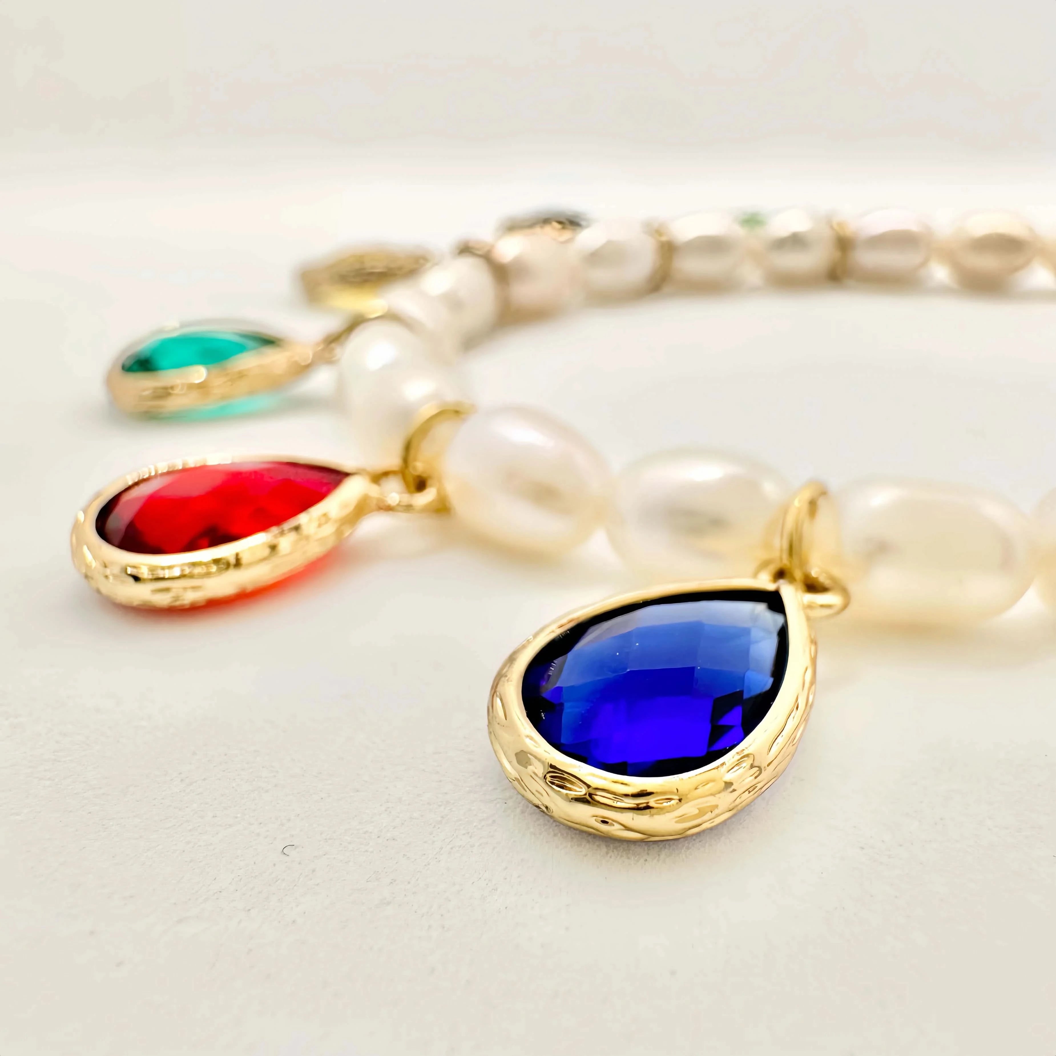 A luxurious pearl necklace and bracelet set displayed on a white background. The necklace is a string of round white pearls, interspersed with gold links and teardrop-shaped colored gemstones in bezel settings. The bracelet mirrors the design with similar colored gems and smaller pearls, both creating a harmonious and opulent look.