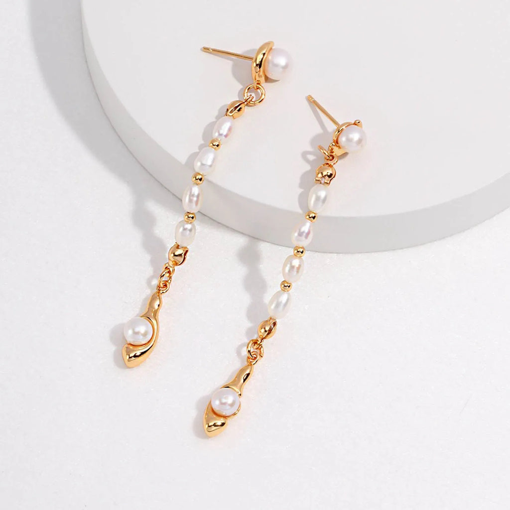 The image shows a pair of gold dangle earrings, each featuring a linear series of small white pearls connected by delicate links. The pearls are evenly spaced, creating an elegant vertical line that culminates in a unique gold charm resembling a stylized figure or a small flame.