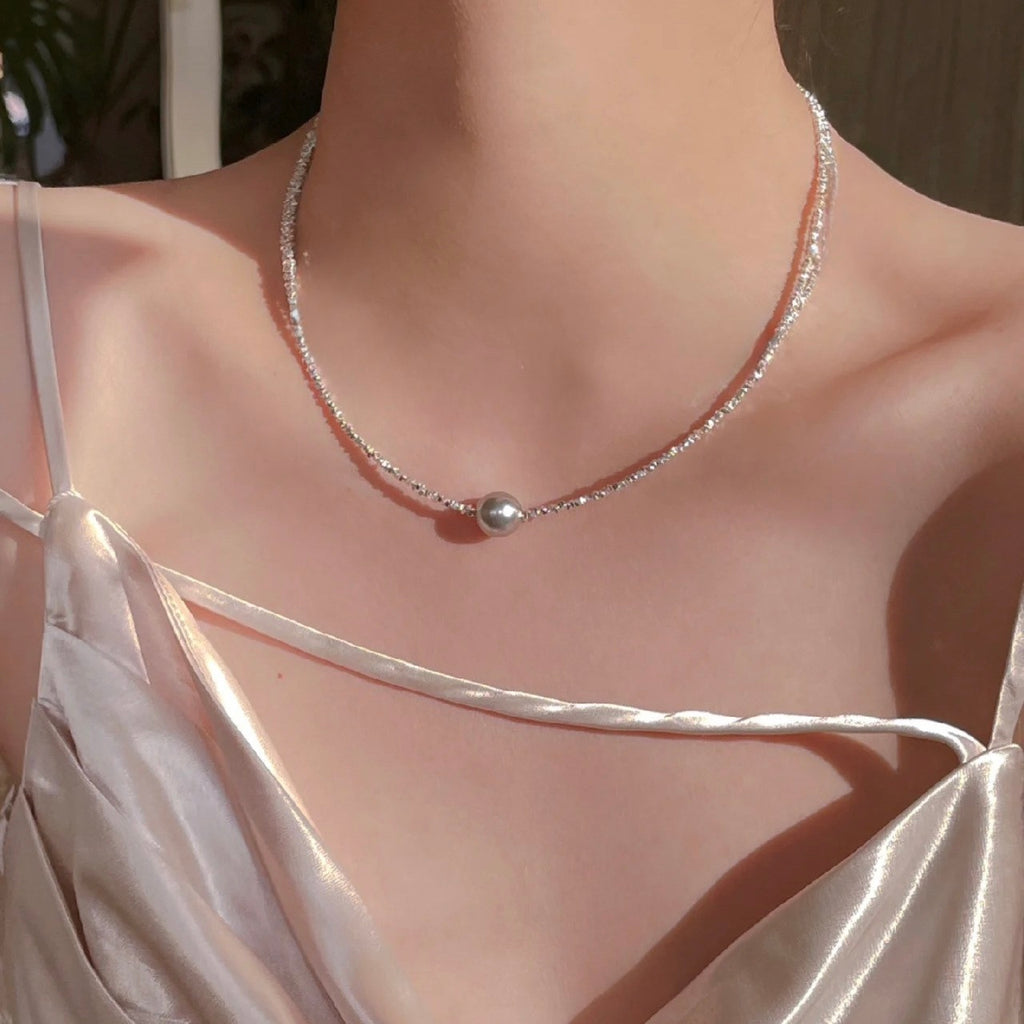 A woman wearing a silk top is adorned with a delicate choker-style necklace, composed of small shimmering beads with a single prominent pearl centered gracefully along her collarbone. The necklace exudes subtle sophistication and highlights the gentle curve of her neck.