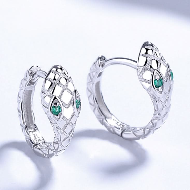 A pair of intricately designed silver snake earrings with a cut-out pattern along the snakes' coiled bodies. Each earring features two small green crystals for eyes, adding a touch of color to the otherwise monochrome palette.