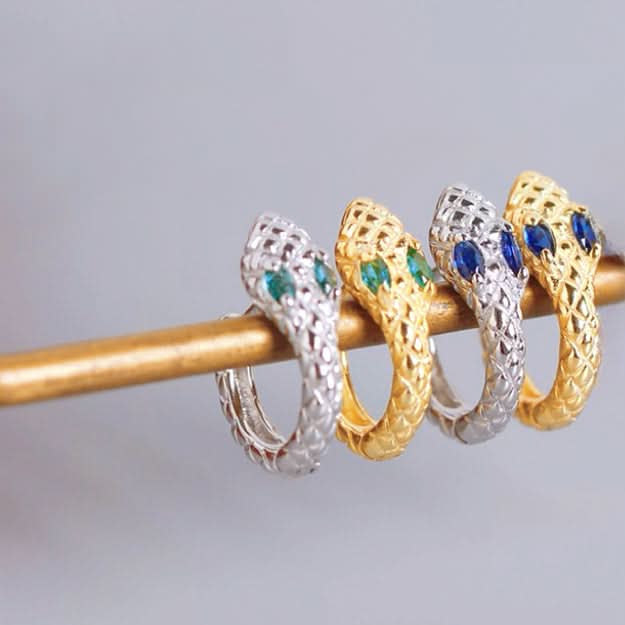 A pair of intricately designed silver snake earrings with a cut-out pattern along the snakes' coiled bodies. Each earring features two small green crystals for eyes, adding a touch of color to the otherwise monochrome palette.