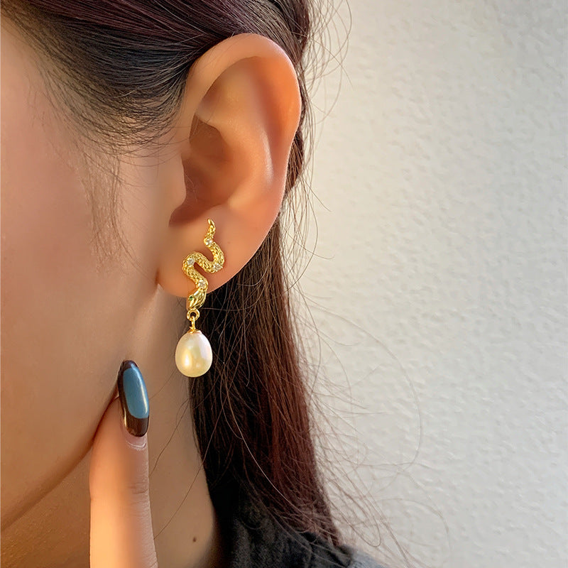 A pair of elegant gold-plated snake earrings, each featuring a sinuous design with textured scales. The snakes are embellished with small diamonds set along their bodies and tiny green crystals for eyes. Each earring is completed with a dangling white pearl at the bottom, adding a classic and sophisticated finish to the piece.