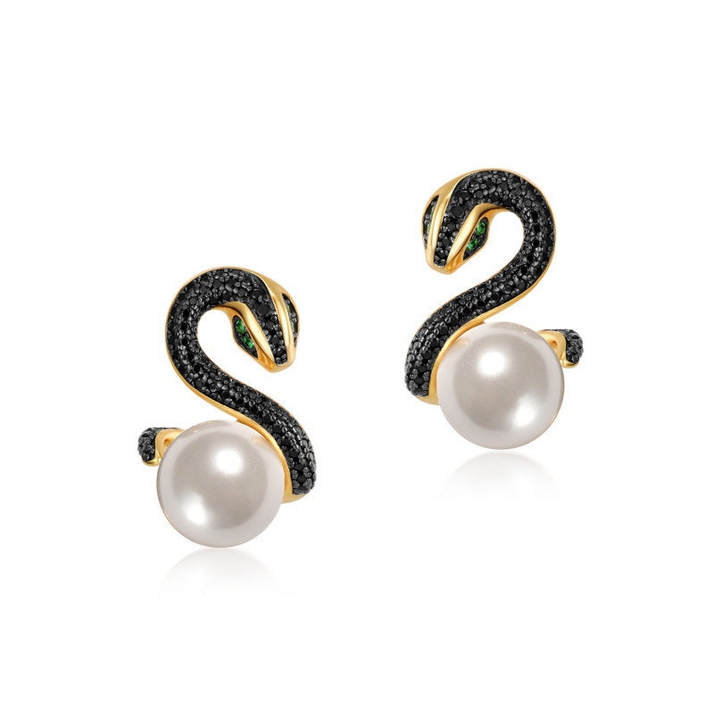 A pair of luxurious gold and black snake earrings, each set with a row of black crystals along the snake's body and featuring vibrant green crystal eyes. The design is anchored by a large white pearl at the bottom, creating a bold and elegant statement piece.