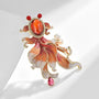 Close-up of orange enamel goldfish brooch with pearl accent and pink teardrop crystal on a light background