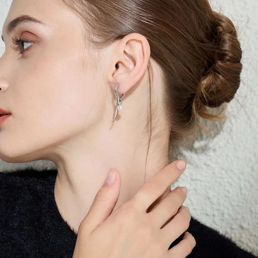 A profile view of a woman wearing a unique silver snake earring. The earring wraps around the edge of her ear, with the snake's head positioned near the top and its body trailing down to end with a small white pearl at the lobe. The snake design includes tiny gemstone accents along its body, providing a subtle sparkle.