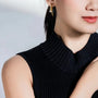 A close-up side view of a woman with a low ponytail, wearing a sophisticated black pleated dress with a high collar. She is adorned with a delicate gold earring that has a small pearl and a crystal detail, giving a subtle hint of elegance to her overall attire.