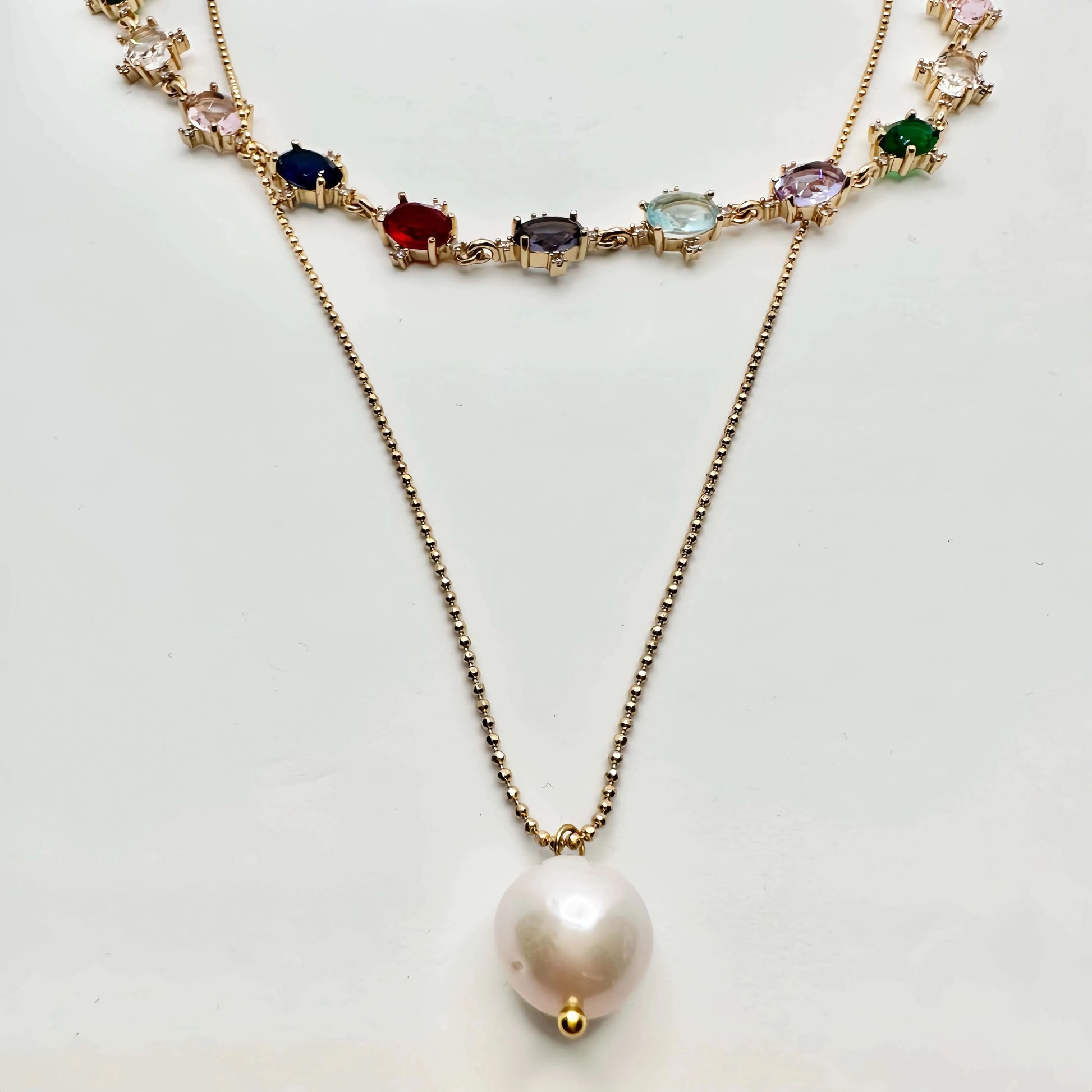 A striking gold necklace laid out on a white surface, featuring a chain of diverse, multicolored gemstones, each set in a gold frame. The necklace transitions into a simple gold chain that culminates in a large, baroque pearl pendant, creating an elegant drop effect. The piece is a blend of classic and contemporary styles.