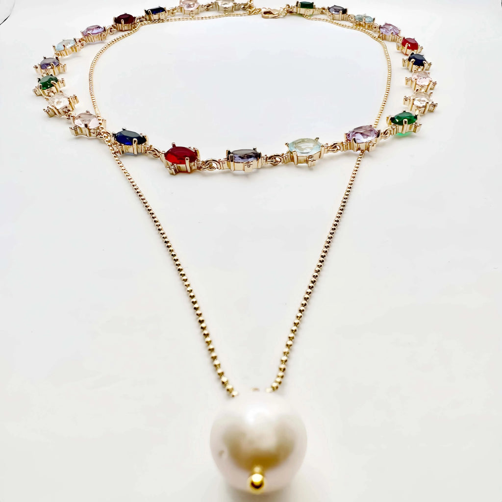 A striking gold necklace laid out on a white surface, featuring a chain of diverse, multicolored gemstones, each set in a gold frame. The necklace transitions into a simple gold chain that culminates in a large, baroque pearl pendant, creating an elegant drop effect. The piece is a blend of classic and contemporary styles.