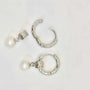 A pair of silver earrings displayed on a white background. Each earring features a textured hoop with an organic, molten-like design, attached to a decorative square bead with intricate patterns. A classic white pearl dangles elegantly from the bead, adding a touch of timeless elegance to the modern design.
