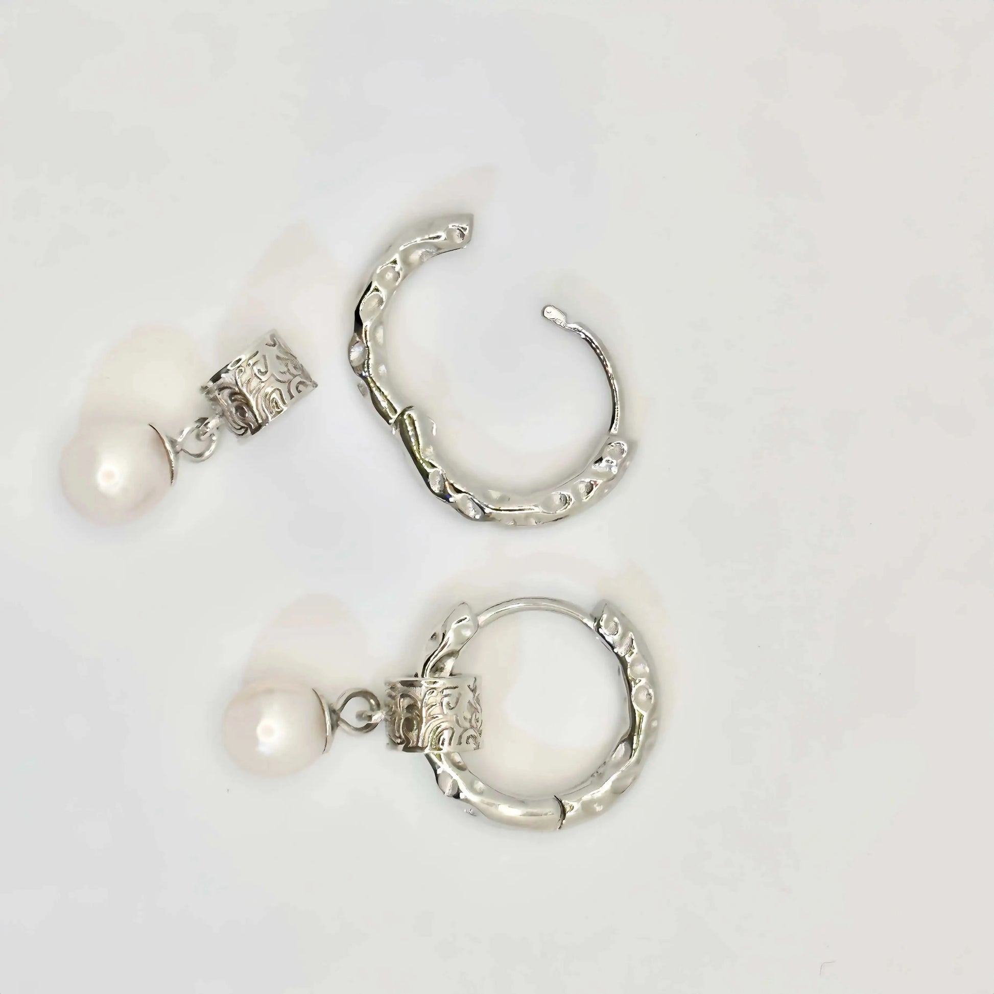 A pair of silver earrings displayed on a white background. Each earring features a textured hoop with an organic, molten-like design, attached to a decorative square bead with intricate patterns. A classic white pearl dangles elegantly from the bead, adding a touch of timeless elegance to the modern design.