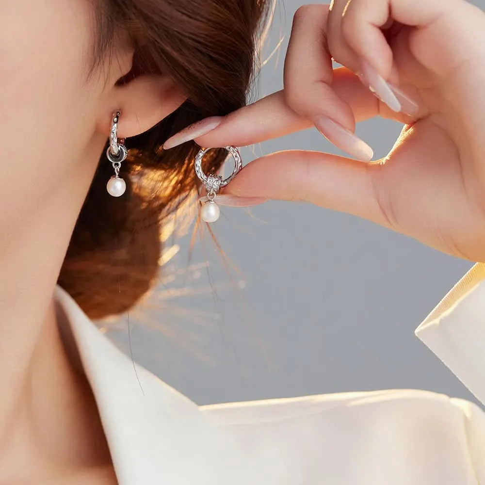 A woman, partially shown, is wearing a sophisticated white blazer and is in the process of adorning her ear with a stylish silver hoop earring that features a small, dangling pearl. The earring's design is both classic and contemporary, highlighting the woman's elegant and refined taste in accessories.