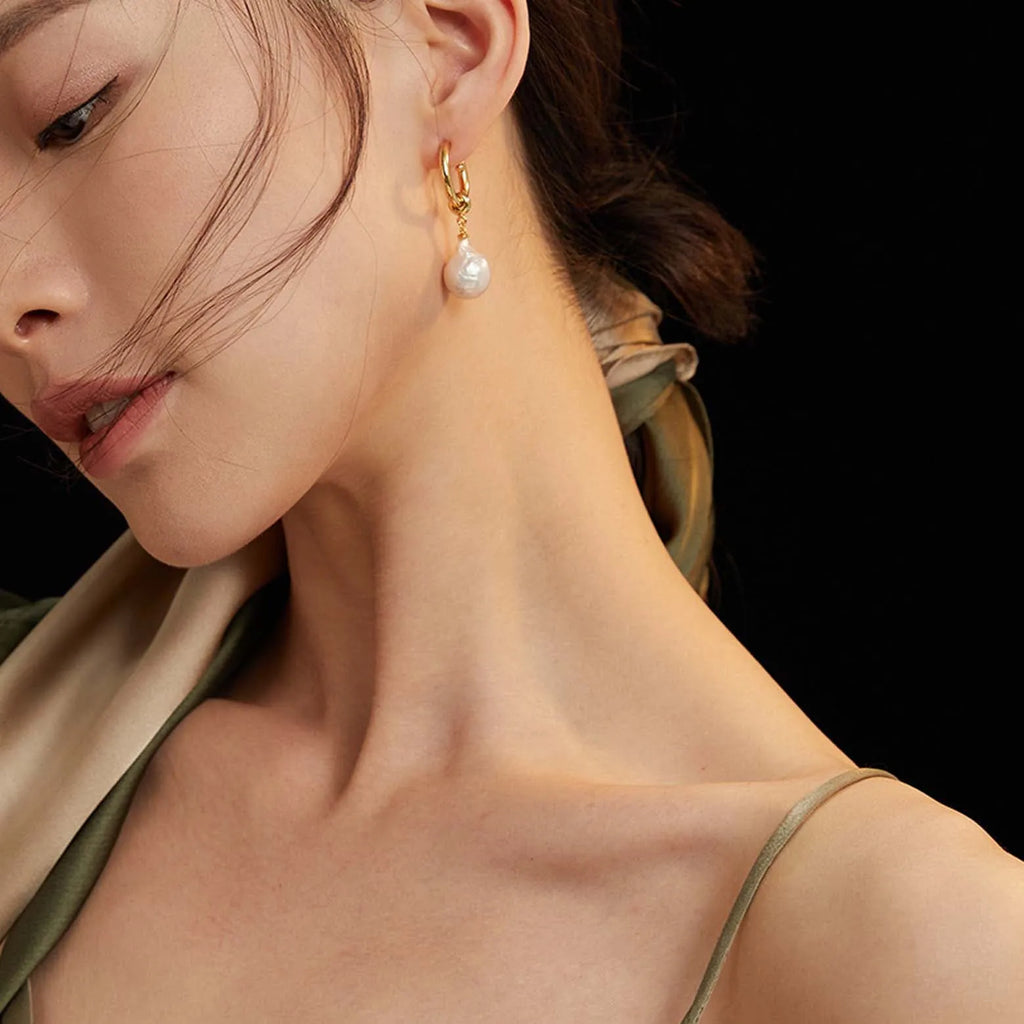 This image captures a woman's profile from the shoulders up, showcasing a gold earring with a white pearl drop. The earring consists of a small gold link attached to a larger, twisted gold element, from which the pearl is suspended, adding a touch of elegance. The pearl's surface is smooth and lustrous, contrasting beautifully with the textured gold.