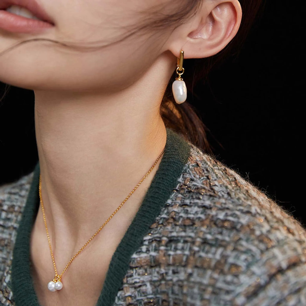  In this image, we see a close-up of a woman's lower face and neckline. She is wearing a gold earring with a single white, elongated pearl drop that exhibits a subtle sheen. The earring's simple design draws attention to the pearl's natural beauty. Complementing the earring is a delicate gold chain necklace with a small, round pearl pendant resting just below the collarbone. The woman's attire includes a dark green top under a textured garment that blends white, black, and hints of metallic threading. 