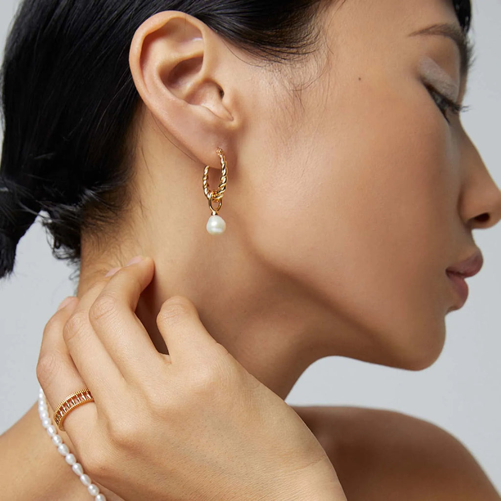 This image captures a woman's profile with a focus on her wearing a gold earring. The earring features a small, twisted gold hoop and a single white pearl drop that adds a touch of elegance. The woman's dark hair is swept back, directing attention to the earring and the smooth line of her neck. She is touching her ear with her hand, which is adorned with a gold ring, complementing the earring. 