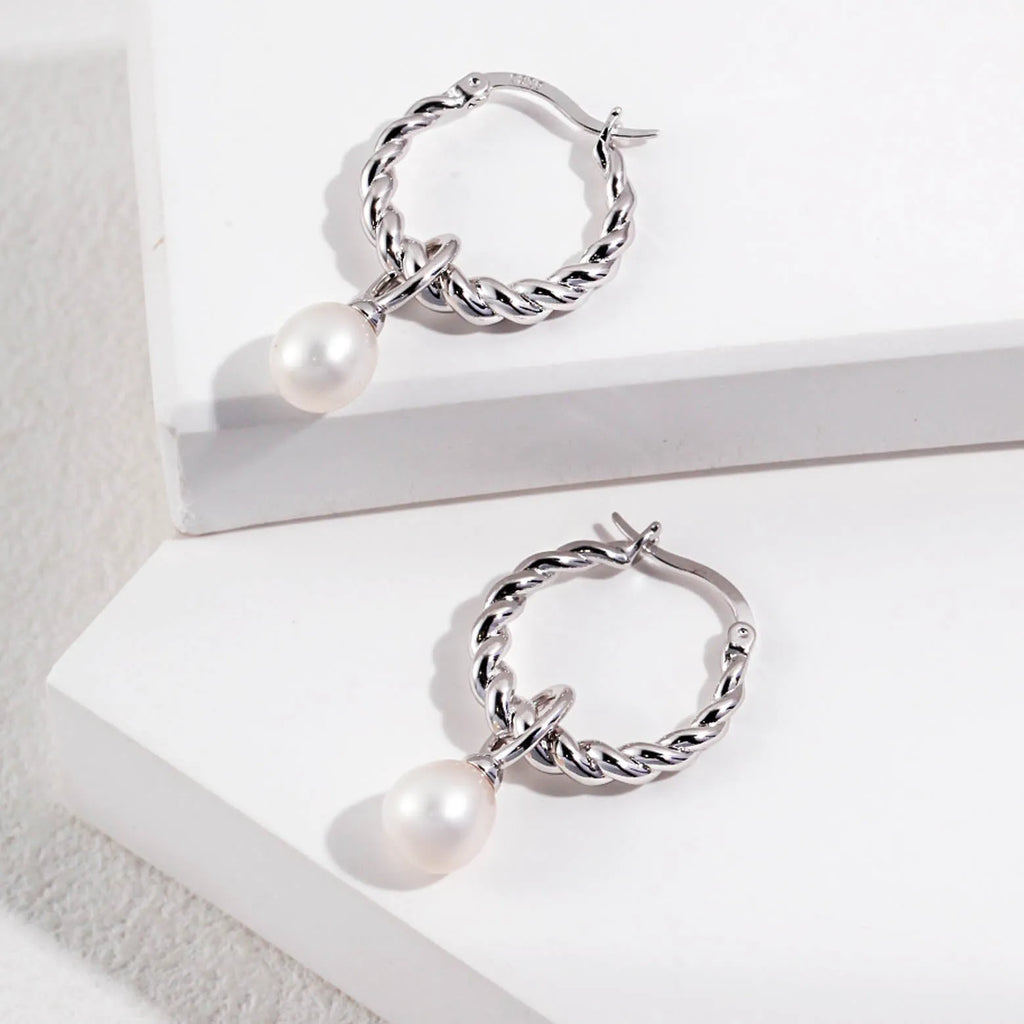 The image showcases a pair of silver hoop earrings with a twisted rope design, providing a sense of texture and depth to the metalwork. Each earring is adorned with a single round pearl pendant, which dangles elegantly below the hoop. The pearls have a soft, lustrous sheen that contrasts beautifully with the polished silver. The earrings are displayed on a white background with subtle shadows, highlighting the shine of the silver and the smoothness of the pearls.