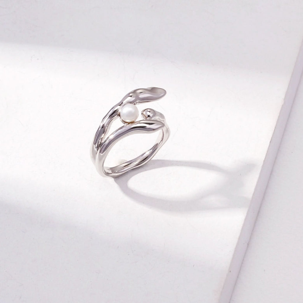This image presents a gold ring with a unique and fluid design, resembling gentle waves that wrap around the finger. At the crest of one of the waves sits a small, round, white pearl that adds an element of classic elegance to the otherwise modern design. The gold has a polished, reflective surface that gleams brightly. The ring is showcased against a white background with an angular shadow, which creates a contrast that highlights the ring's dynamic curves and the pearl's soft luster.
