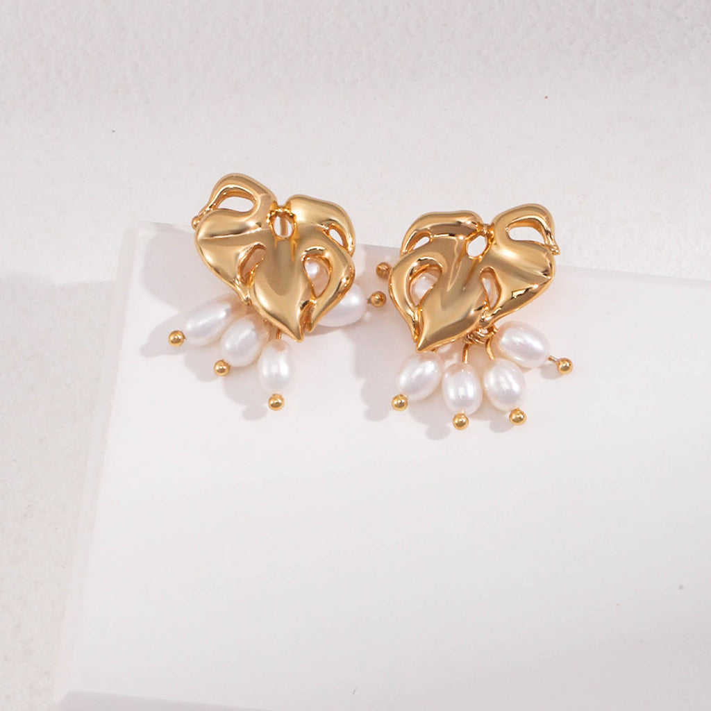 The image showcases a pair of gold stud earrings, each with an artistic and organic heart-shaped design featuring openwork details. From each earring, three white oval pearls dangle, giving the piece a sense of movement and elegance. The pearls are attached to the gold hearts with small, round gold beads that add an extra detail to the design.