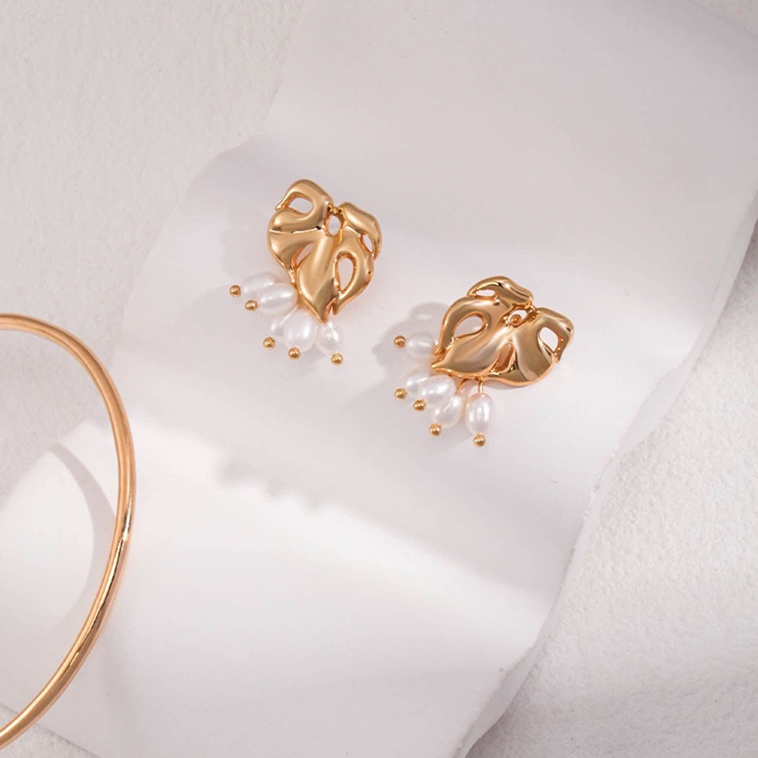 The image shows a pair of gold stud earrings, each featuring an abstract, openwork heart-shaped design. The hearts have a polished, sculptural quality, with smooth curves and holes that create a dynamic pattern. Suspended from each heart are three white oval pearls, attached by small gold links that end in round gold beads. 