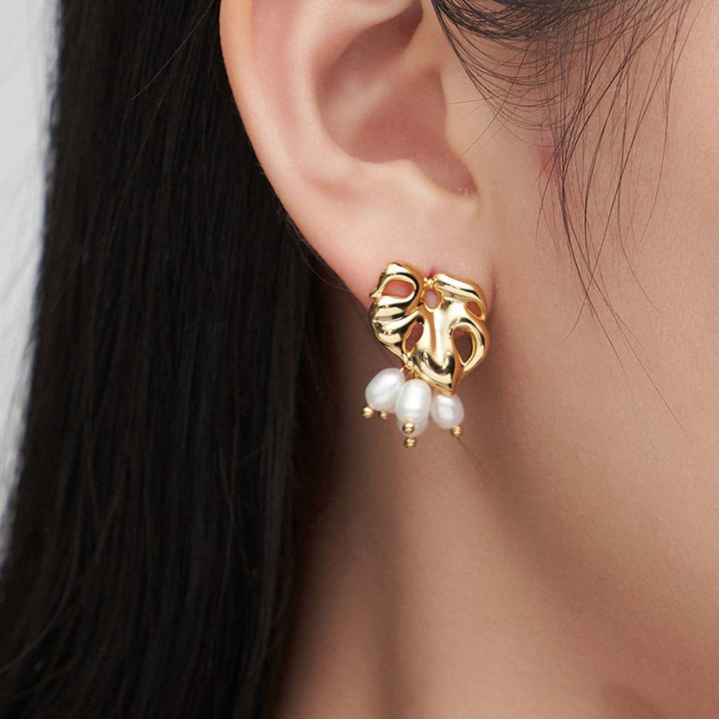 The image features a close-up of a woman's ear adorned with a gold earring. The earring has a heart-shaped design with cut-out details and a polished surface that reflects light. Three white pearls dangle from the bottom of the heart, each pearl separated by a tiny gold bead, adding movement and a classic touch to the piece. The woman's dark hair provides a contrasting backdrop that accentuates the gold's warm tones and the pearls' soft luster.