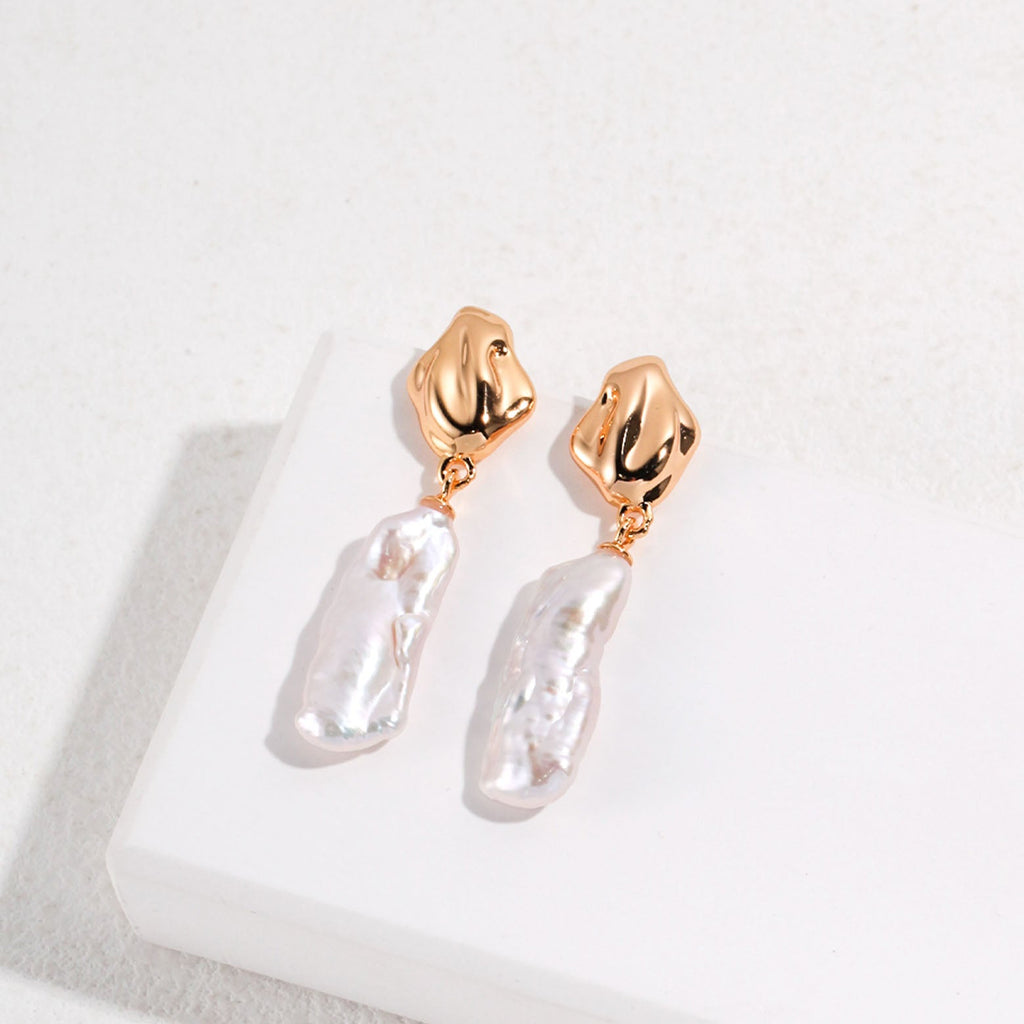 The image displays a pair of gold dangle earrings, each consisting of a polished, gold-toned abstract stud and a large, irregularly shaped baroque pearl pendant. The pearls have a natural luster with an uneven surface that reflects light in various directions, creating a beautiful sheen. The organic forms of the pearls contrast with the smooth, sculpted appearance of the gold studs. These earrings are designed to make a statement, blending contemporary style with the timeless elegance of pearls.
