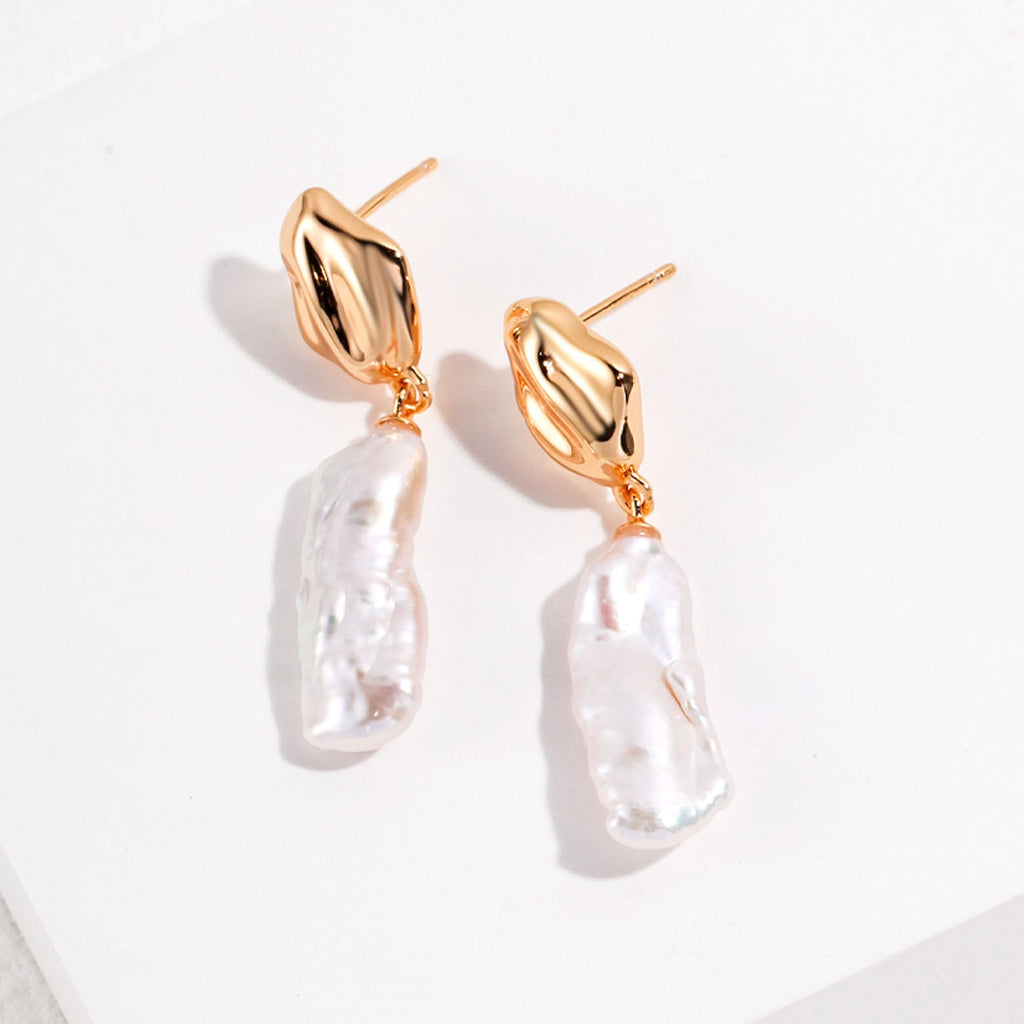 The image displays a pair of gold dangle earrings, each consisting of a polished, gold-toned abstract stud and a large, irregularly shaped baroque pearl pendant. The pearls have a natural luster with an uneven surface that reflects light in various directions, creating a beautiful sheen. The organic forms of the pearls contrast with the smooth, sculpted appearance of the gold studs. These earrings are designed to make a statement, blending contemporary style with the timeless elegance of pearls.