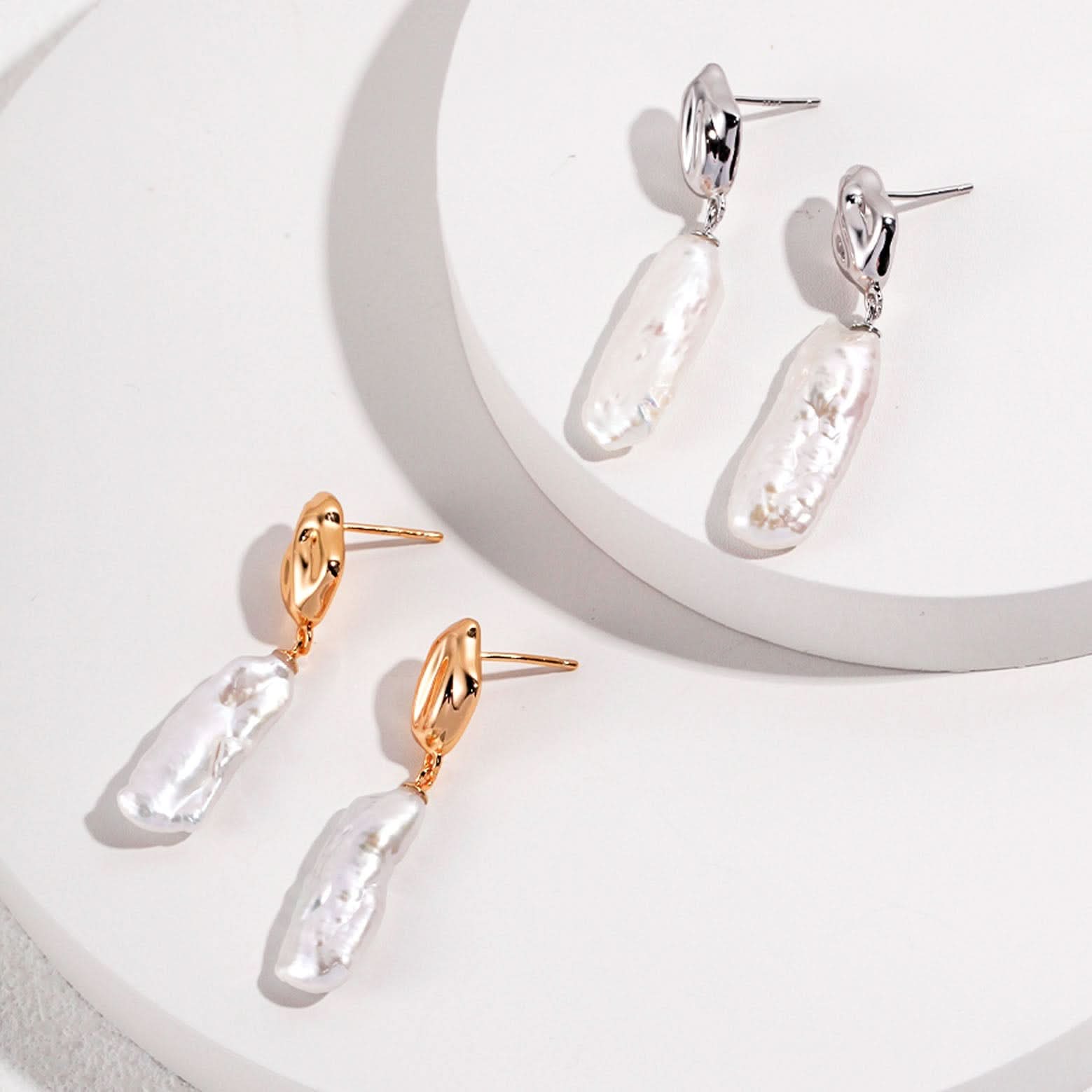 The image displays a pair of gold dangle earrings, each consisting of a polished, gold-toned abstract stud and a large, irregularly shaped baroque pearl pendant. The pearls have a natural luster with an uneven surface that reflects light in various directions, creating a beautiful sheen. The organic forms of the pearls contrast with the smooth, sculpted appearance of the gold studs. These earrings are designed to make a statement, blending contemporary style with the timeless elegance of pearls.